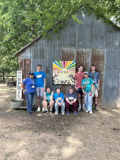 Large family group standing and sitting together in front of a rustic barn with trees overhead