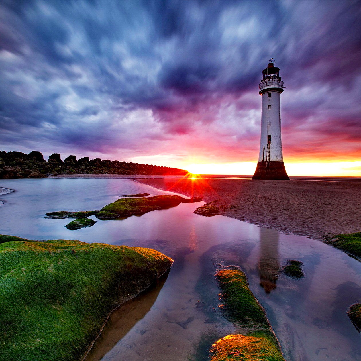 White lighthouse stands on beach at sunset with colorful sky reflected in water pools surrounded by mossy rocks