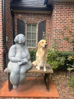 Golden retriever sitting on a bench next to a gray statue of a woman reading, in front of a brick house