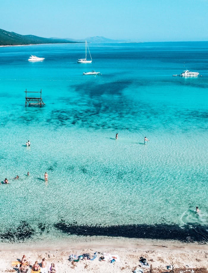 Turquoise beach lagoon with swimmers, boats, and a pebbly shore