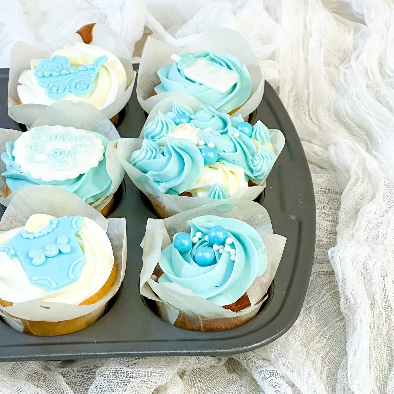 Six cupcakes with blue swirled frosting in white paper liners on a dark baking tray