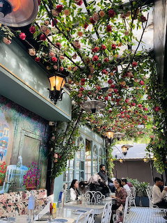 Outdoor cafe terrace with roses and vines hanging from overhead trellis, lanterns, and seated diners