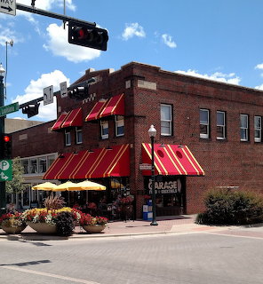 Brick corner cafe with red awnings, patio umbrellas, and flowers at a sunny intersection