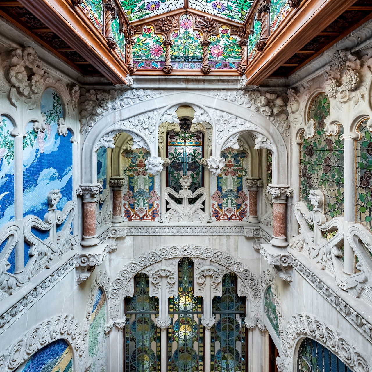 Ornate multi-level palace hall with white carved columns, arches, decorative railings, and colorful glass ceiling