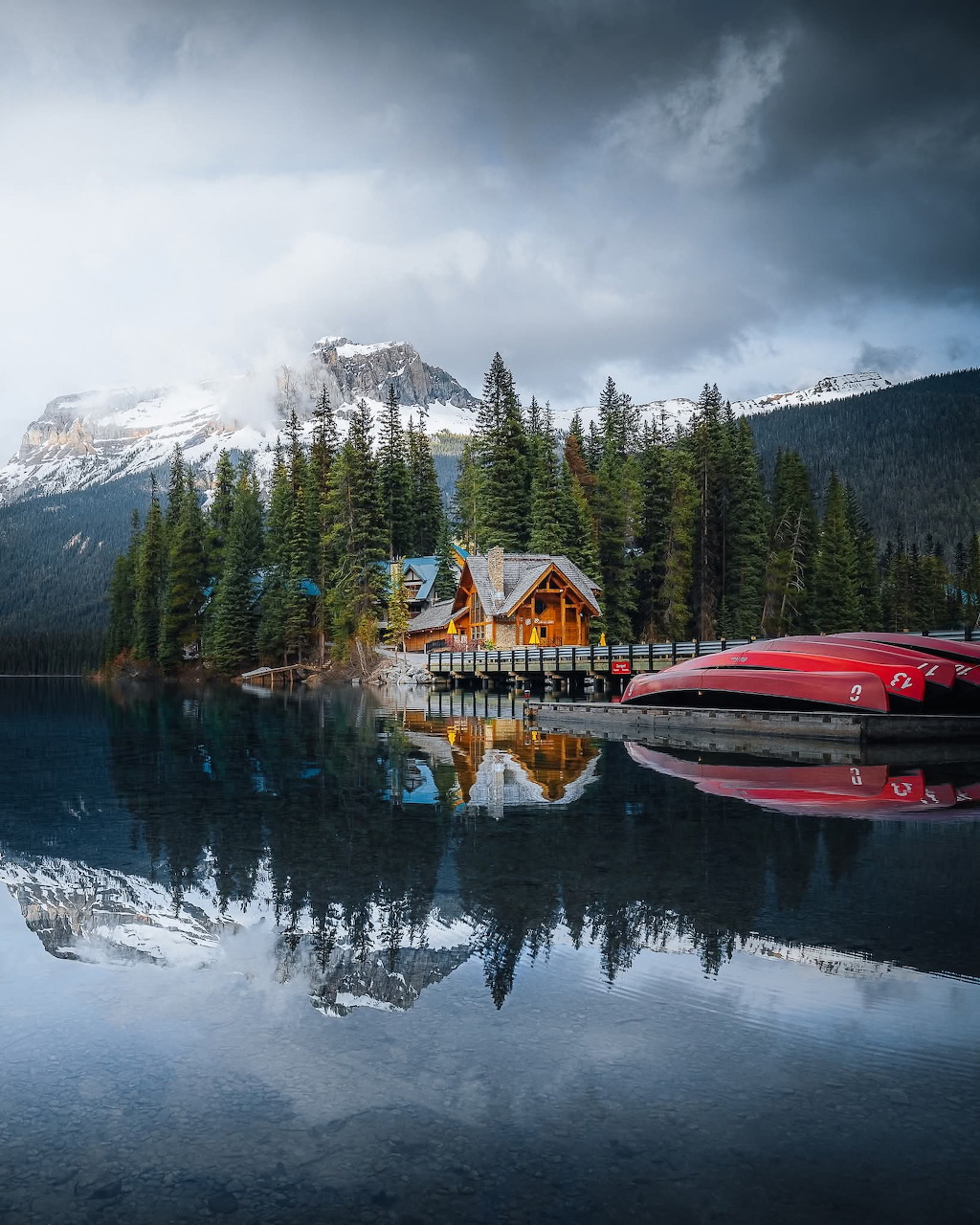 Wooden lodge reflected in a calm alpine lake with pine trees and snow-capped mountains in background