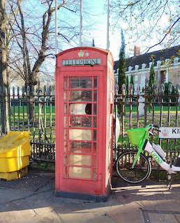 Red British telephone box on a sidewalk with a green bicycle leaning nearby and iron railings behind