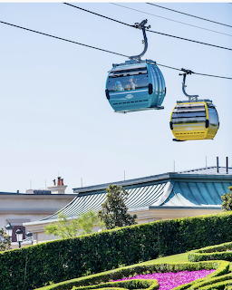 Blue and yellow Disney Skyliner gondola cabins on cables above garden landscape