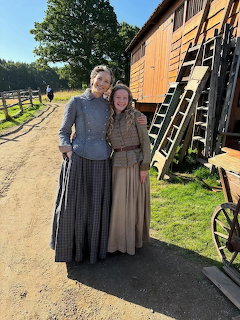 Two women in historical costumes posing together outdoors on a film set with wooden buildings