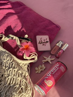 Pink flatlay with burgundy towel, woven bag, sunscreen, flower, and beach accessories on pink background
