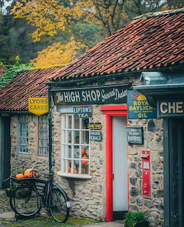 Old stone cottage shop with red door, bicycle parked outside, clay tile roof, autumn trees in background