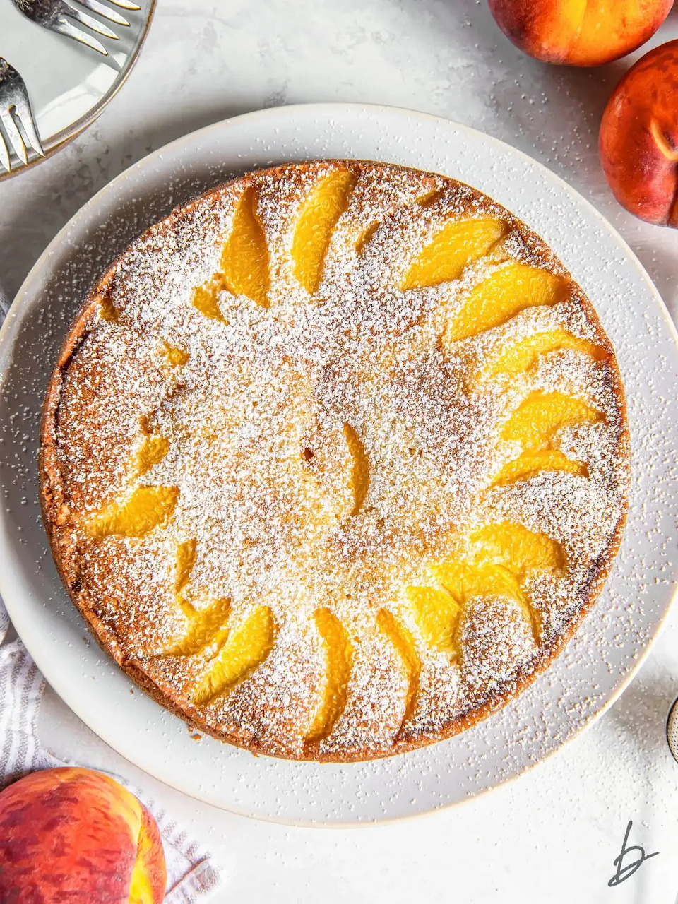 Round cake with peach slices arranged in a sunburst pattern, dusted with powdered sugar on a white plate