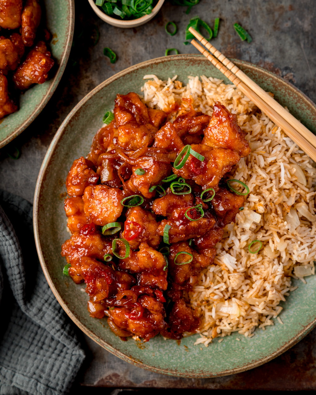 Ceramic plate of spicy glazed chicken in red sauce with steamed rice, green onion garnish, and chopsticks