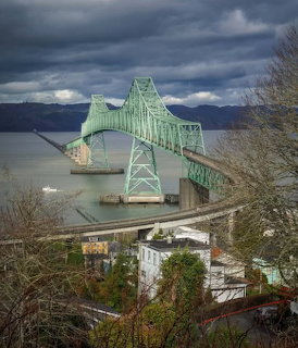 Green steel truss Astoria-Megler Bridge crossing the Columbia River with cloudy skies and town buildings below