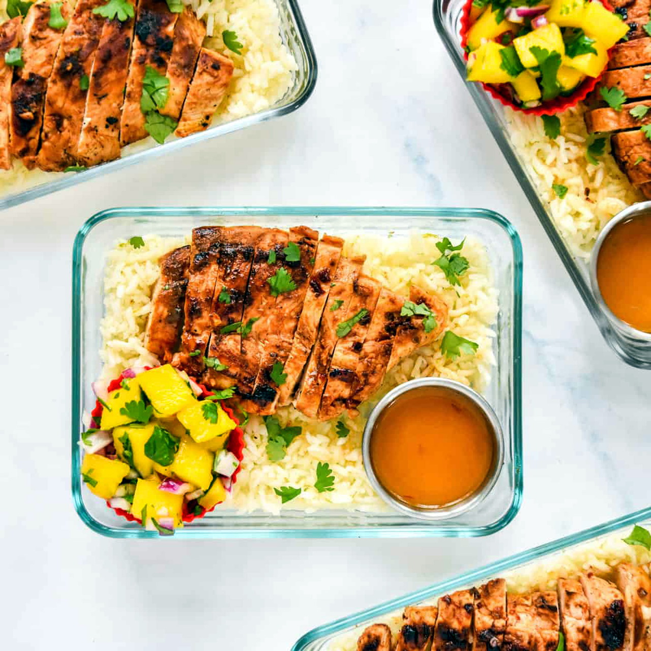 Meal prep containers with grilled chicken, rice, mango salsa, and a small cup of sauce