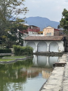 White arched pavilion reflected in a calm pond with buildings and mountains behind it