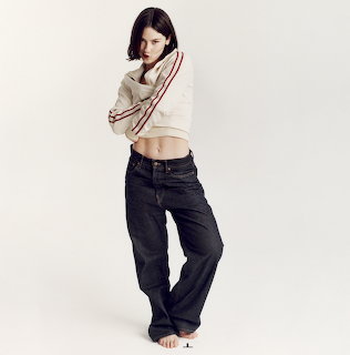 Full-body studio portrait of Gracie Abrams wearing a cropped sweater and dark jeans on a white background