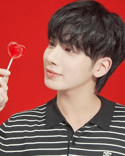 Young man with black hair holding a red heart lollipop in striped polo on red background