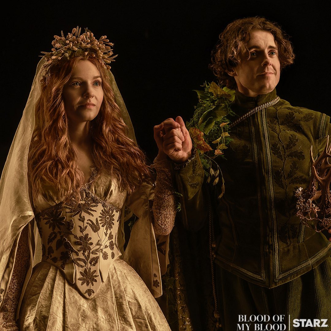 Bride in golden gown with ornate crown beside man in green medieval jacket