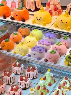 Colorful cute fruit-shaped cakes with kawaii faces displayed in a bakery glass case