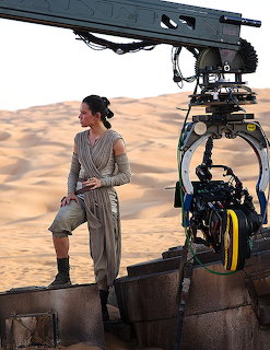 Woman in beige costume standing on platform beside large film camera in desert landscape