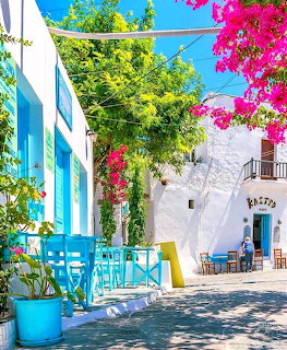 Colorful Greek village street with white buildings, blue furniture, pink bougainvillea flowers, and green trees