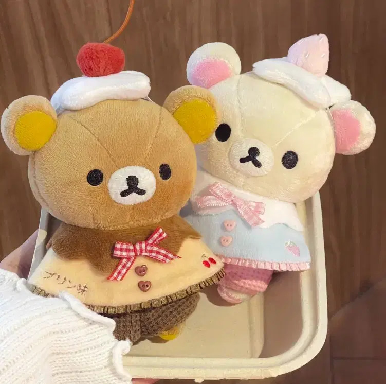 Two Rilakkuma plush bears in costumes, one with cherry hat and pancake outfit, other in pink apron with cake