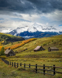 Wooden cabins on grassy hillside with snow-covered mountain peaks and storm clouds above