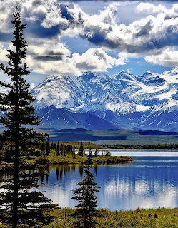 Alaska mountain range reflected in calm lake with pine trees in foreground under cloudy sky