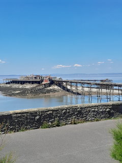 Historic Victorian pier with buildings extending over water, connected by walkway to rocky shore under blue sky