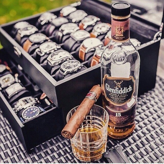 Glenfiddich whisky bottle with glass and cigar beside a case of luxury watches