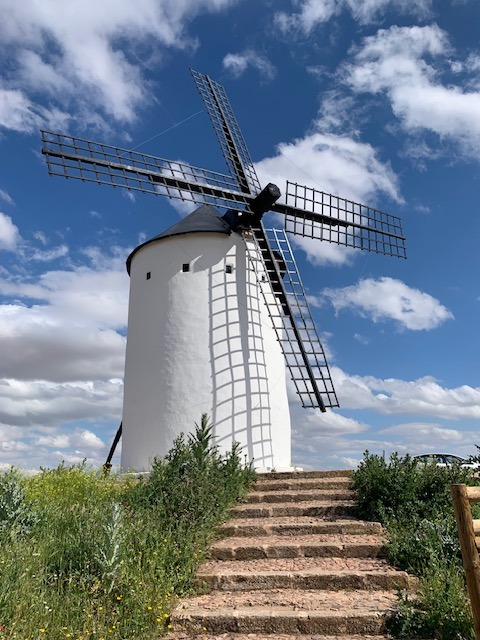 White traditional windmill with lattice blades on a green hilltop under a blue cloudy sky with stone steps