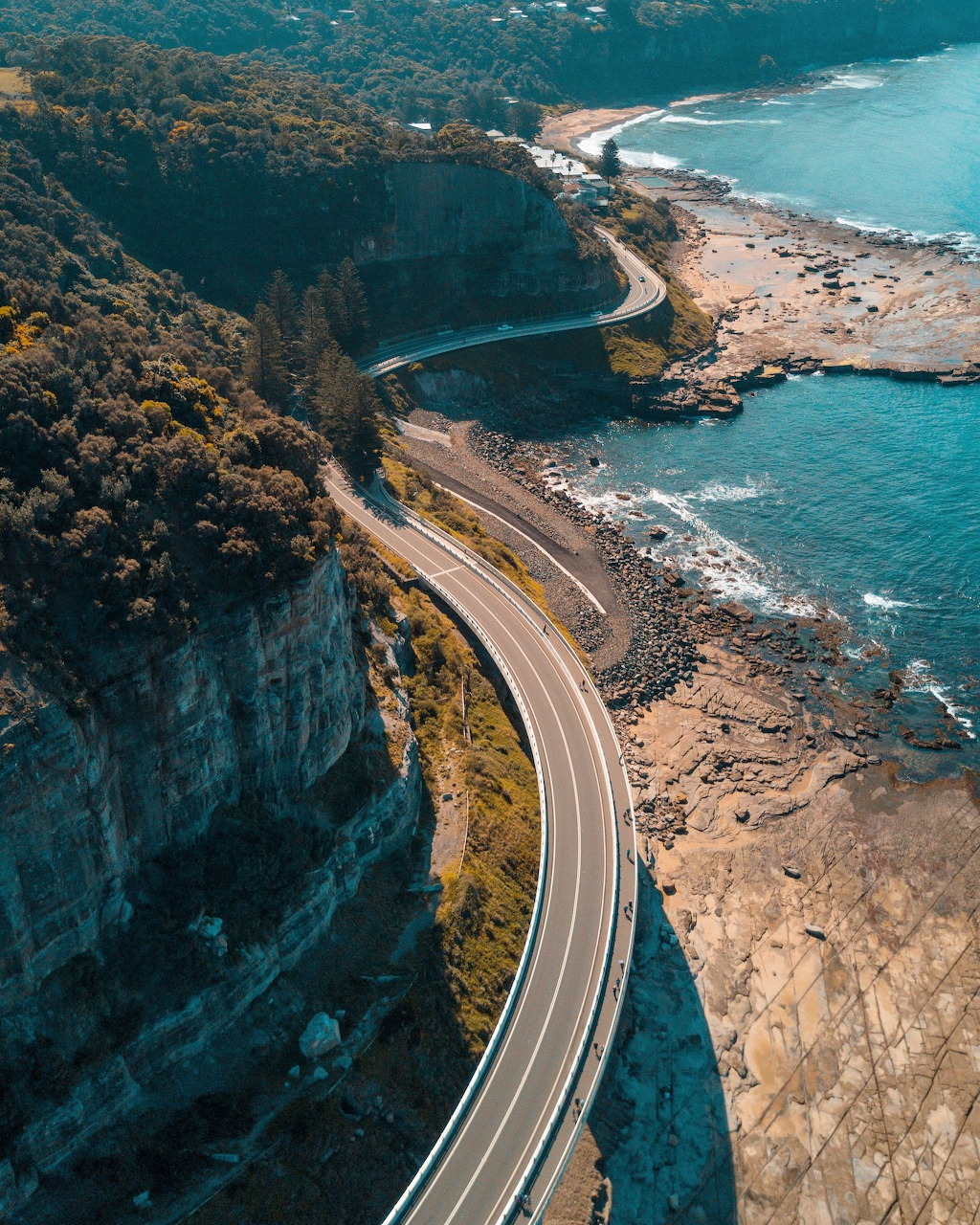 Aerial view of curved coastal road between forested cliffs and turquoise ocean