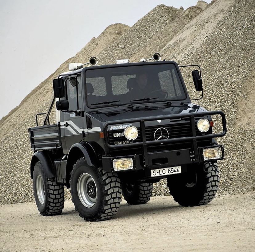 Black Mercedes Unimog off-road truck parked on sandy ground in front of gravel hills