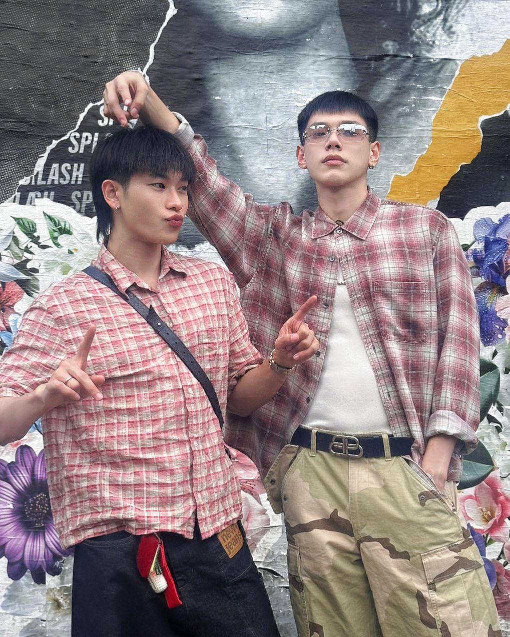 Two young men in pink plaid shirts posing together against artistic floral background