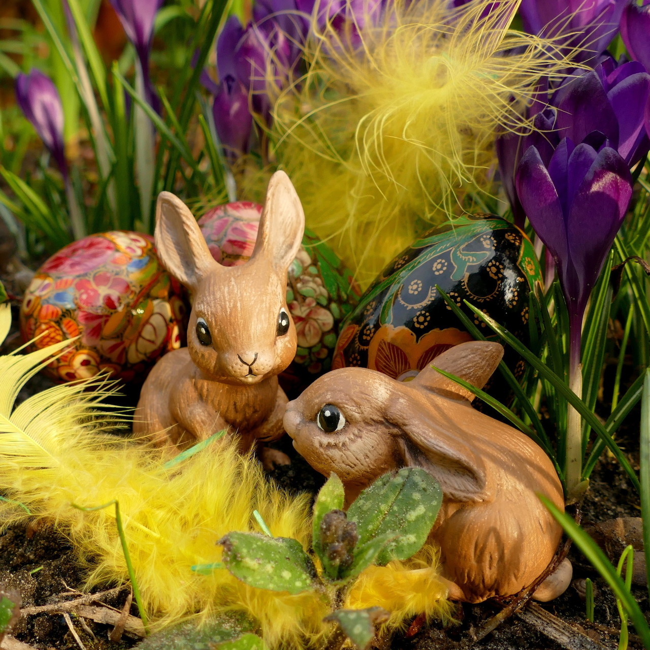 Ceramic Easter bunny figurines with painted eggs, yellow feathers, and purple crocus flowers in spring arrangement