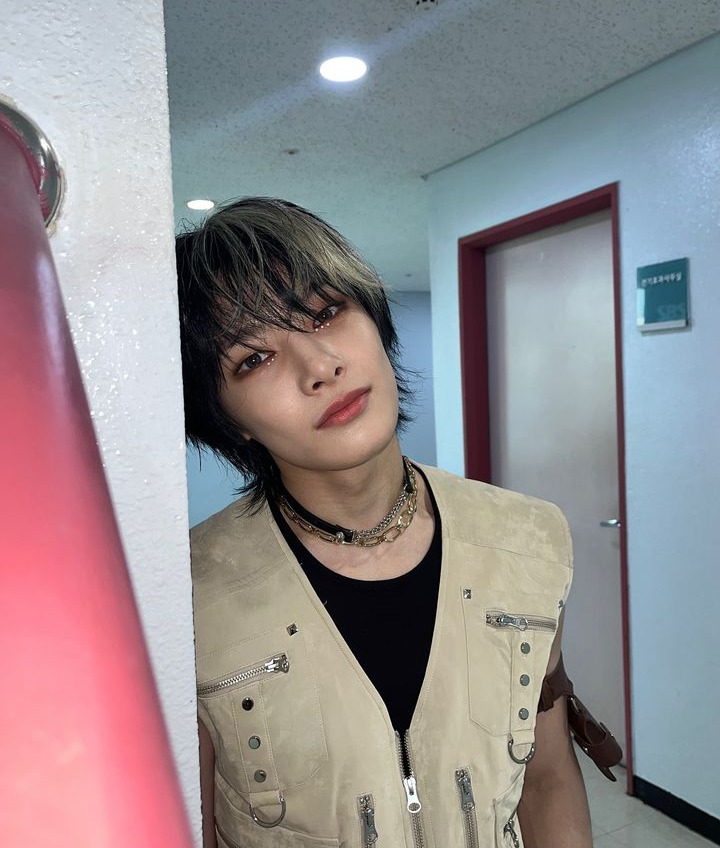 Young man with layered hair wearing a beige vest and chain necklace posing in a hallway