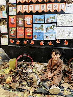 Young child sitting on floor with pine cones and autumn crafts in front of kindergarten art display