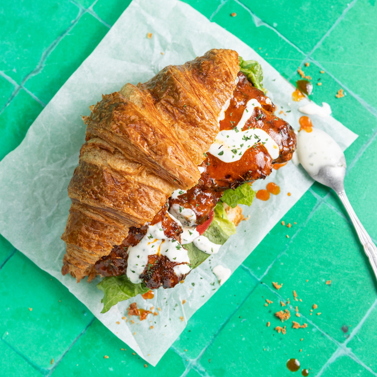 Croissant sandwich with meat, greens, and cream on parchment over turquoise tiles