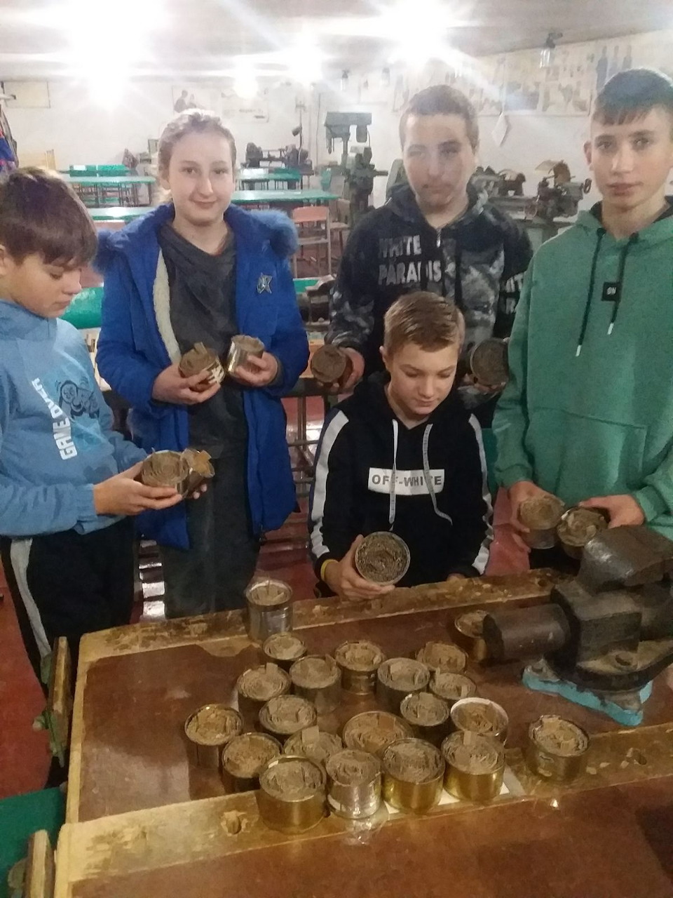 Five teenagers examining metal cans and parts at a workbench in a factory or workshop