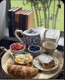Breakfast tray with teapot, pastries, fruit, and eggs beside books by a window overlooking a green garden