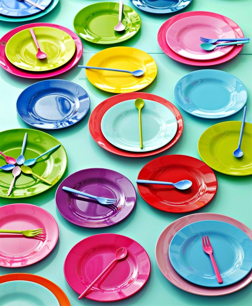 Assortment of colorful plastic plates, spoons, and forks on a mint green background