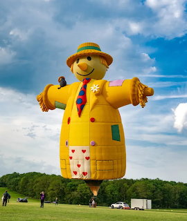 Scarecrow-shaped hot air balloon on a grassy field under a blue cloudy sky