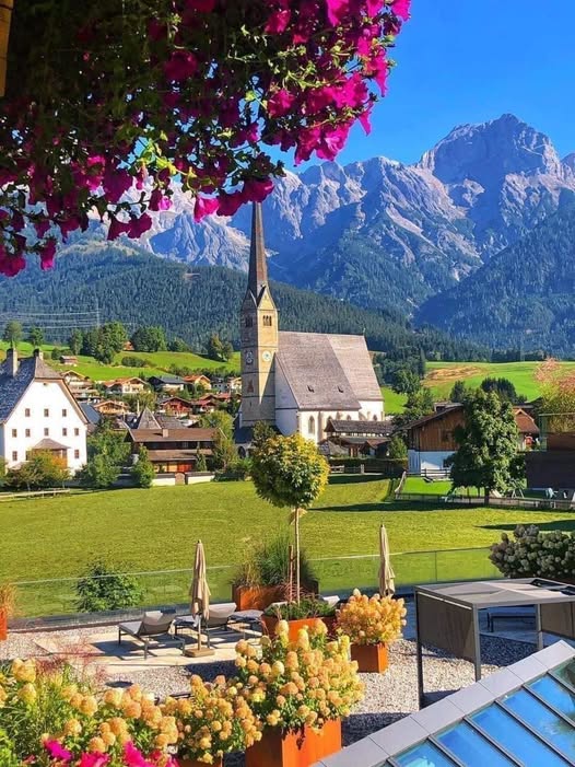Alpine village with white church steeple, green meadows, colorful flowers, and rocky mountain peaks in background