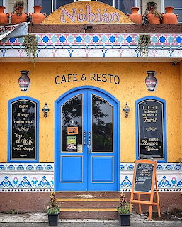 Colorful cafe entrance with blue ornate doors, yellow facade, decorative tiles, hanging lanterns, and chalkboard signs