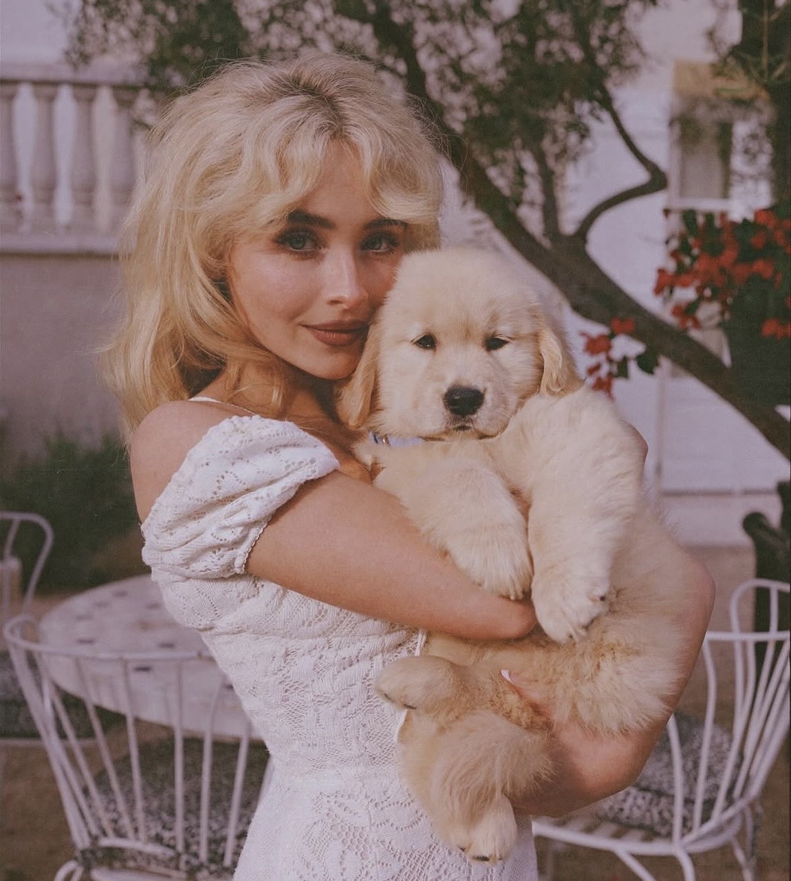 Woman with blonde hair in white lace dress holding white fluffy puppy in garden
