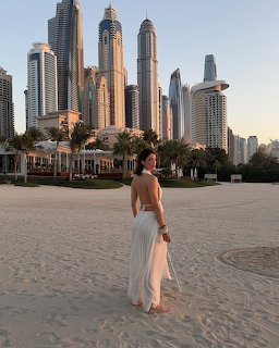 Woman in white dress standing on sandy beach with Dubai Marina skyscrapers in the background at sunset