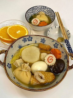 Bowl of Japanese oden containing fish cakes, boiled egg, daikon, and vegetables with orange slices nearby