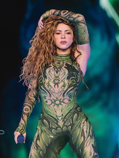Shakira singing on stage wearing an elaborate green sequined outfit with long wavy blonde hair
