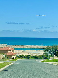 Road descending toward a blue ocean with houses, sandy shore, and breakwater in Perth, Western Australia