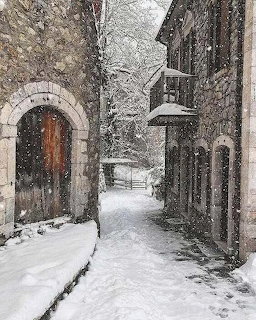 Snow-covered narrow alley between stone buildings with an arched wooden door and falling snowflakes